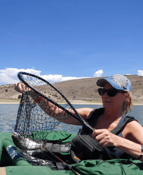 A woman in a blue cap holds a fishing net with a fish while kayaking on a sunny day.