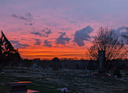 A vibrant sunset casts orange and pink hues over a cemetery, with gravestones and trees silhouetted against the sky.