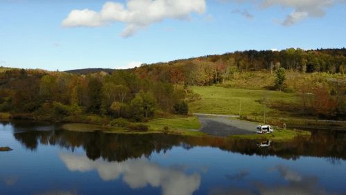 A serene landscape featuring a calm lake reflecting trees and clouds, with a parked vehicle by the shore.