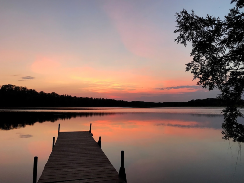 A serene lake at sunset, with a wooden dock extending into calm waters reflecting vibrant pink and orange hues.
