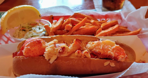 A lobster roll with chunks of lobster on a bun, served with sweet potato fries and a lemon wedge.
