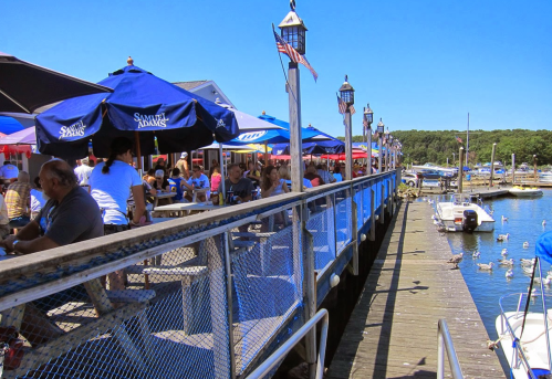 A bustling waterfront pier with diners under umbrellas, boats docked, and a clear blue sky.