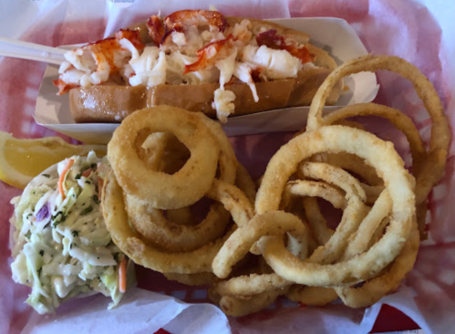 A lobster roll topped with lobster meat, served with onion rings and a side of coleslaw on a red and white checkered tray.