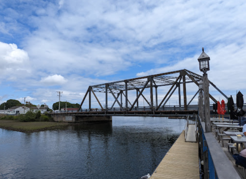 A metal bridge spans a calm river under a partly cloudy sky, with a wooden dock and seating area in the foreground.