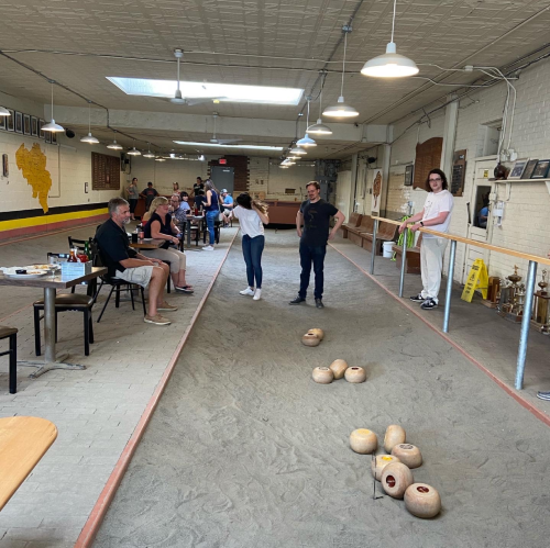 A bowling alley with players and spectators, featuring a sandy lane and wooden pins in a casual setting.