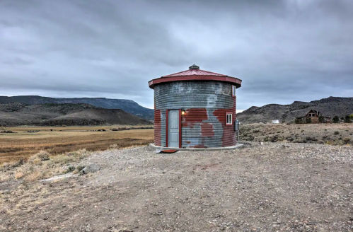 A round, corrugated metal building stands alone in a barren landscape under a cloudy sky, surrounded by mountains.