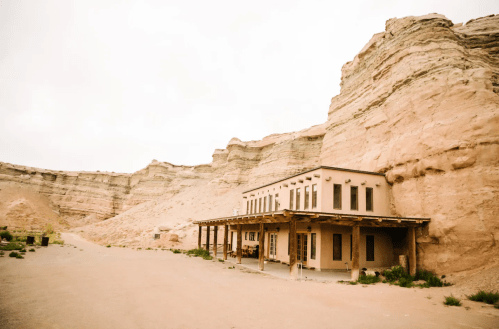 A rustic building sits at the base of a rocky cliff in a desert landscape, surrounded by sparse vegetation.