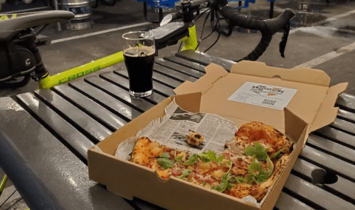 A half-eaten pizza in a box on a table, with a glass of dark beer and a bicycle in the background.