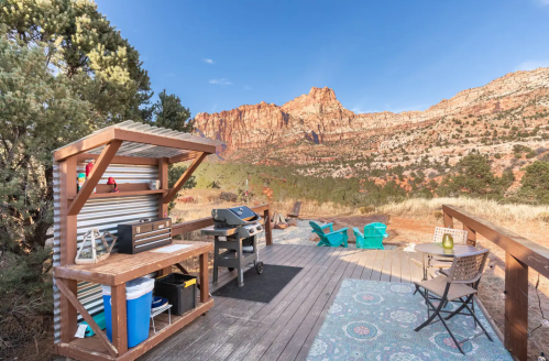 Outdoor kitchen area with a grill and seating, surrounded by mountains and trees under a clear blue sky.