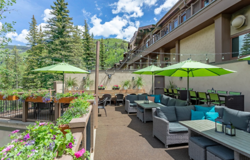 A cozy outdoor patio with green umbrellas, comfortable seating, and vibrant flowers, set against a mountain backdrop.
