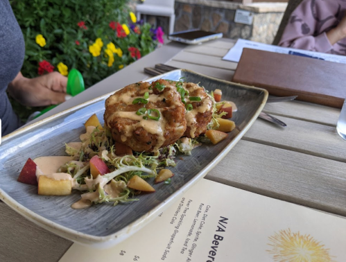 A plate of crispy crab cakes topped with sauce, served on a bed of slaw and diced apples, with colorful flowers in the background.