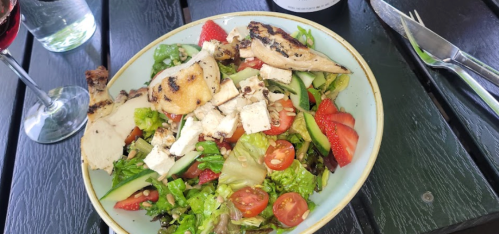A colorful salad with grilled chicken, strawberries, tomatoes, and greens, served with a glass of red wine.