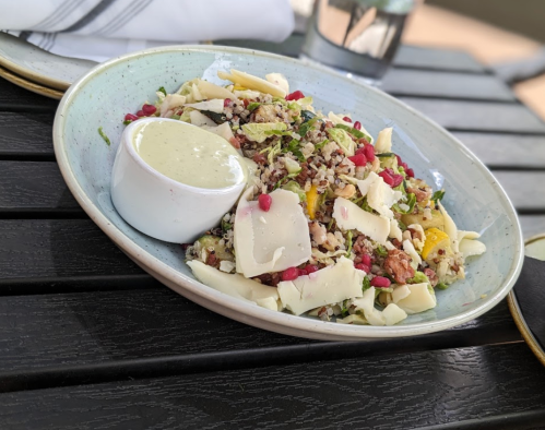A colorful salad with quinoa, pomegranate, and shaved cheese, served with a creamy dressing on the side.