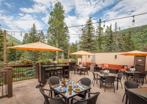 Outdoor dining area with tables, umbrellas, and scenic views of trees and mountains under a partly cloudy sky.