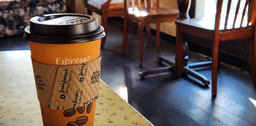A coffee cup labeled "Espresso" sits on a table in a cozy café with wooden chairs and soft lighting.