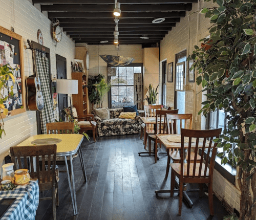 Cozy café interior with wooden tables, chairs, plants, and a sofa, featuring warm lighting and a relaxed atmosphere.