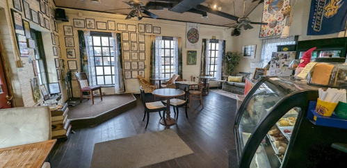 Cozy café interior with framed artwork, seating areas, and a display case, featuring large windows and wooden floors.