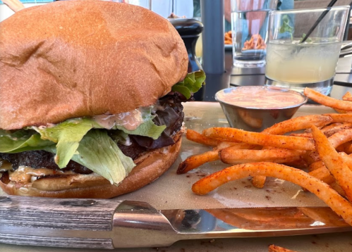 A juicy burger with lettuce and sauce, served with crispy fries on a plate, with drinks in the background.