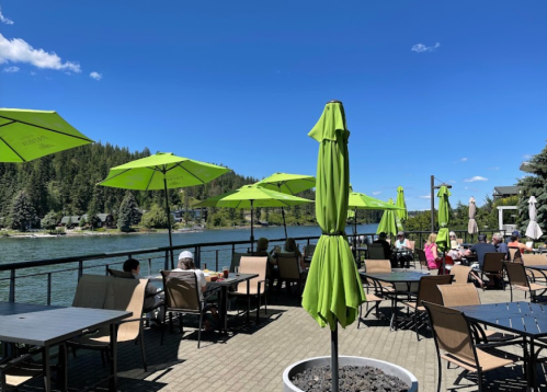 Outdoor dining area by a lake, featuring green umbrellas and tables, with people enjoying the sunny day.
