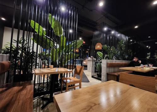 A modern restaurant interior with wooden tables, plants, and black dividers, featuring a person sitting at a table.
