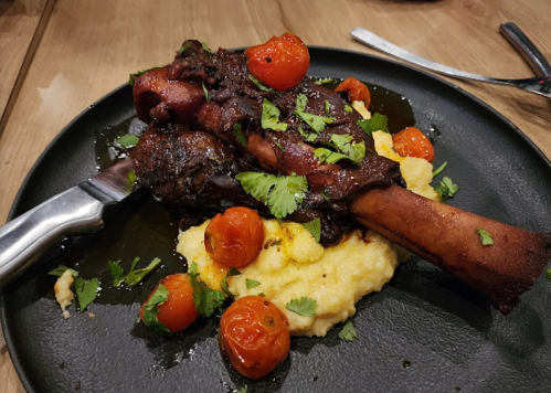 A plate featuring a large, glazed meat shank on creamy polenta, garnished with cherry tomatoes and fresh herbs.