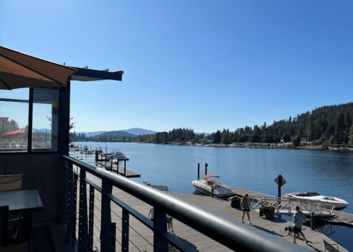 A scenic view of a lake with boats, surrounded by trees and mountains, from a deck with a railing. Clear blue sky.