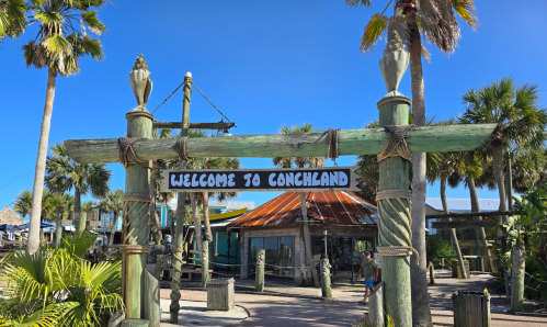 Sign reading "Welcome to Conchland" at a tropical location with palm trees and a blue sky.