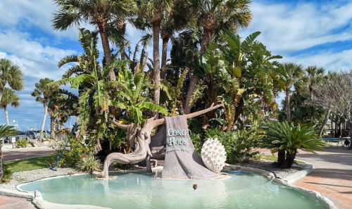 A tropical scene featuring a fountain with a sign reading "Conch House" surrounded by palm trees and lush greenery.