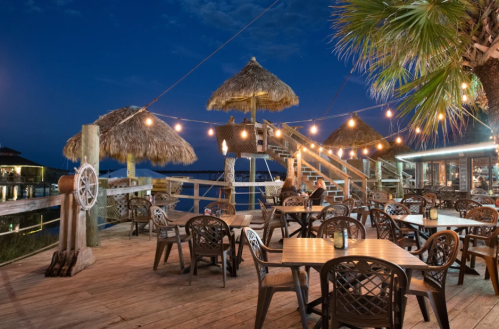 A waterfront dining area with thatched umbrellas, string lights, and wooden tables at dusk.