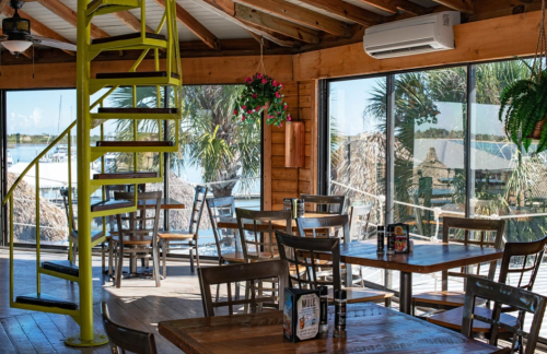 Bright, airy restaurant interior with wooden tables, chairs, and a spiral staircase, overlooking a waterfront view.