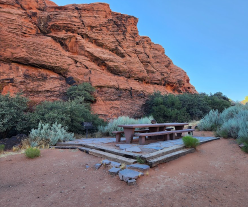 A picnic area with a stone table and benches, surrounded by red rock formations and desert vegetation.