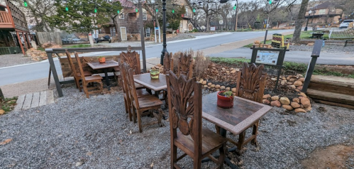 Outdoor dining area with wooden tables and chairs, surrounded by gravel and greenery, near a quiet street.