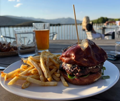 A gourmet burger with a pretzel bun, served with fries, on a table by a lake with mountains in the background.