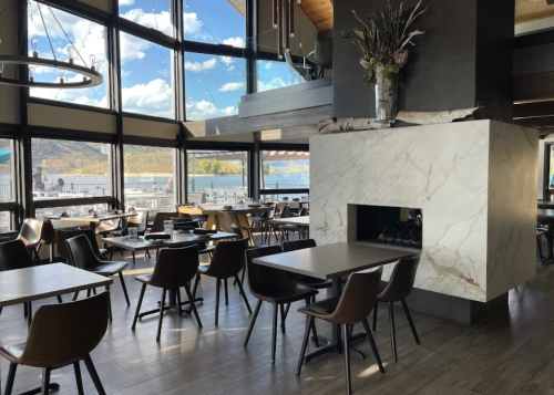 Modern restaurant interior with large windows, wooden tables, and a marble fireplace, overlooking a scenic view.