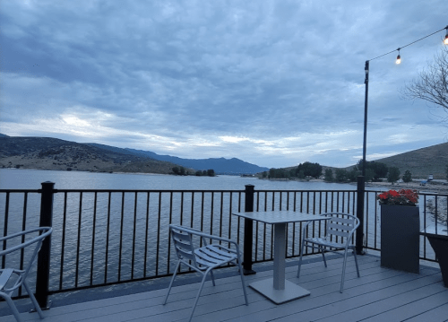 A serene lakeside view from a deck, featuring a table and chairs, with mountains and cloudy skies in the background.