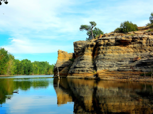 A serene landscape featuring a rocky cliff reflected in calm water, surrounded by lush greenery and a clear blue sky.