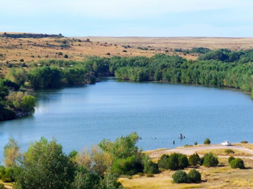 A serene river surrounded by green trees and grassy hills under a clear blue sky.