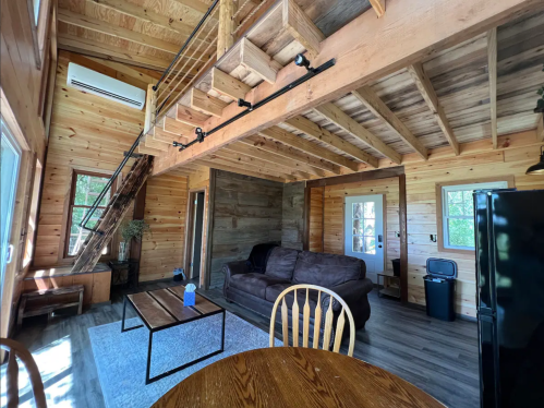 Cozy wooden cabin interior featuring a living area, dining table, and lofted staircase. Natural light fills the space.