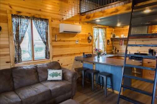 Cozy wooden interior of a tiny home featuring a sofa, kitchen, and lofted sleeping area with large windows.