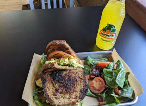 A toasted sandwich with greens, tomatoes, and a bottle of Jarritos soda on a tray next to a side salad.