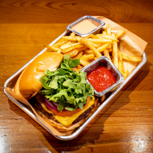 A burger with cheese, lettuce, and onion, served with fries and dipping sauces in a metal tray on a wooden table.