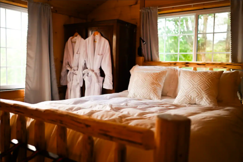 Cozy bedroom with a wooden bed, white bedding, and two bathrobes hanging by the window. Natural light fills the room.