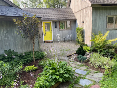 A charming garden path leads to a bright yellow door, surrounded by greenery and two white chairs.