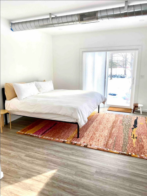 Bright bedroom with a large bed, white bedding, a colorful rug, and a sliding glass door leading to an outdoor view.
