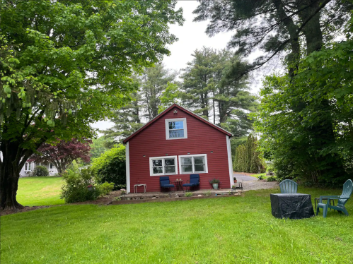 A red house surrounded by green trees and grass, with a patio and chairs in the yard.