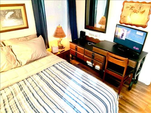 Cozy bedroom with a striped bedspread, wooden dresser, lamp, and a TV on a desk. Natural light from a window.