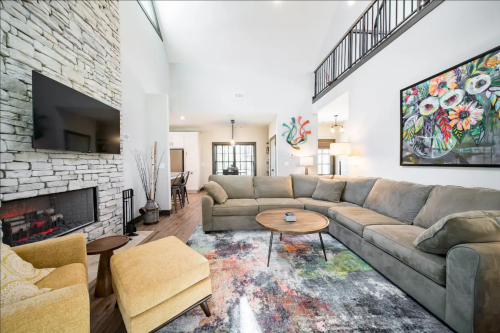 Cozy living room with a stone fireplace, large sectional sofa, colorful rug, and vibrant artwork on the walls.