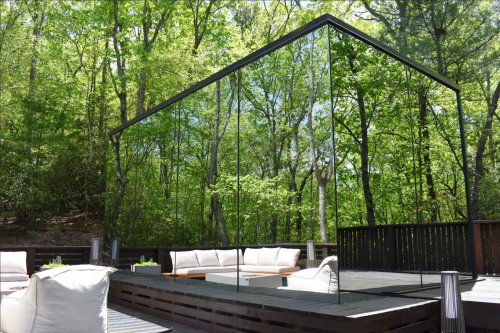 A modern glass structure reflects lush greenery, surrounded by a wooden deck and outdoor seating.