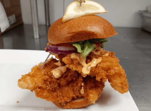 A crispy fried chicken sandwich topped with lettuce, tomato, onion, and a lemon wedge on a toasted bun.