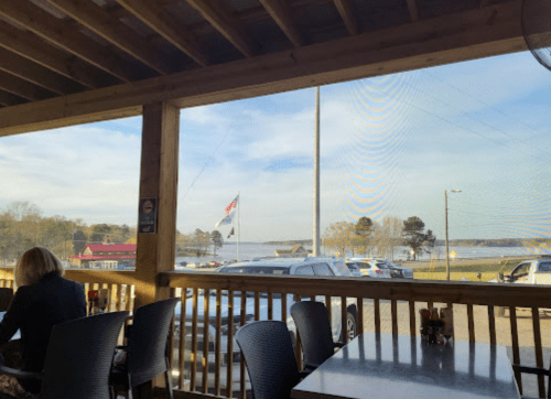 View from a restaurant porch showing a lake, parked cars, and an American flag in the distance.